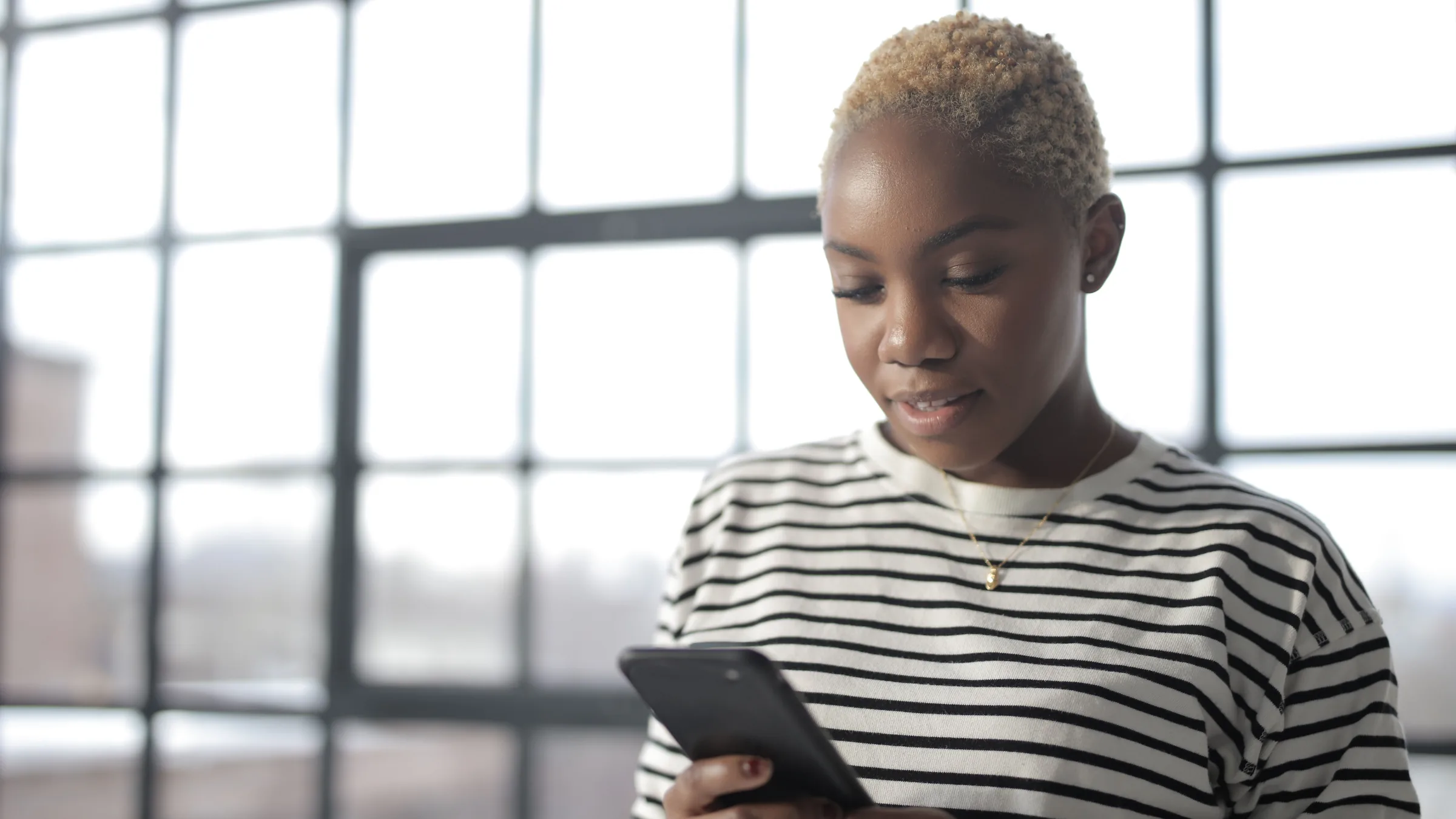A young woman with short blonde hair in a striped top looking at her phone beside a large window