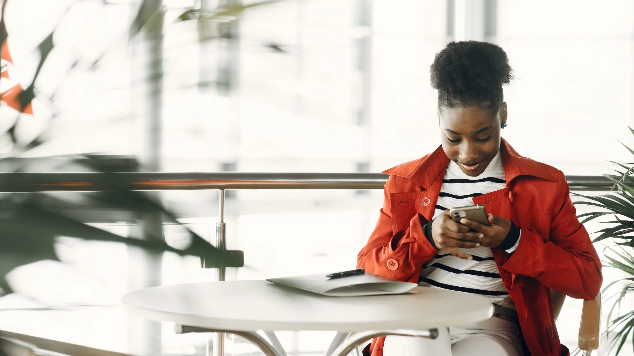 A young woman in a red coat smiling at her phone at a table in a bright space